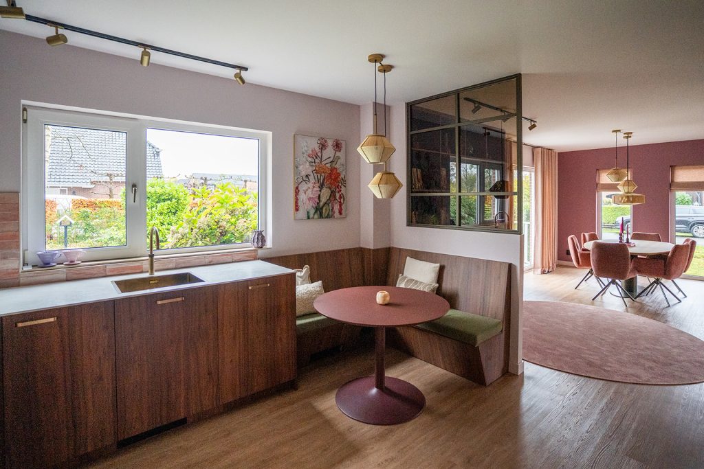 Maatwerk keuken met geïntegreerde eethoek, zitbank, warme houttinten, roze accenten en stalen binnenraam in een woonhuis in Winschoten, ontworpen door Studio Vogt en gerealiseerd door A&F Interieurbouw.