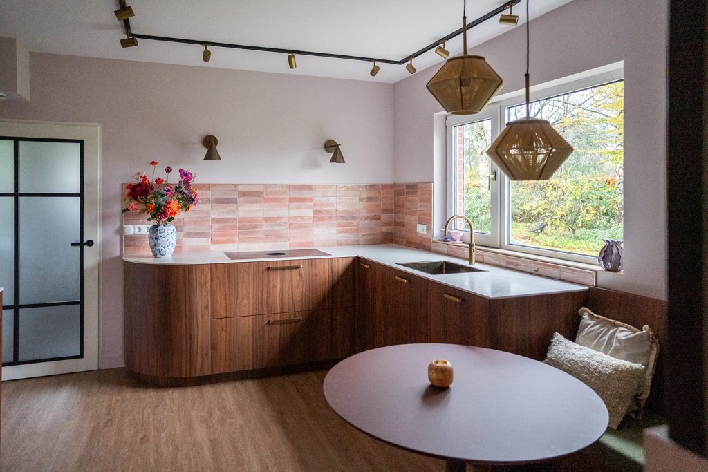 Maatwerk keuken met warme houttinten, afgeronde vormen, roze wandtegels en geïntegreerde eethoek in een woonhuis in Winschoten, ontworpen door Studio Vogt en gerealiseerd door A&F Interieurbouw. Groningen